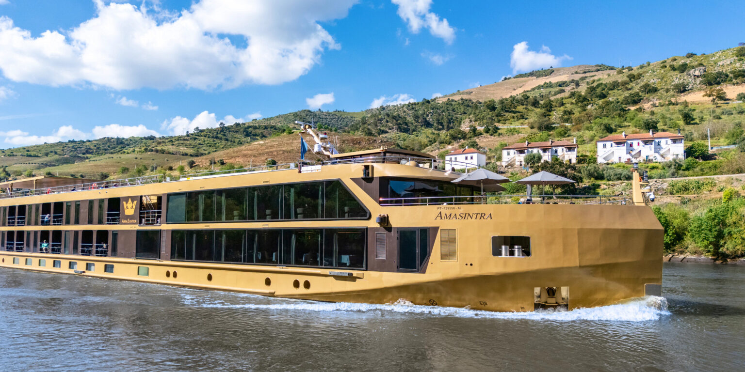 A large, modern riverboat cruising on a sunny day with hills in the background.