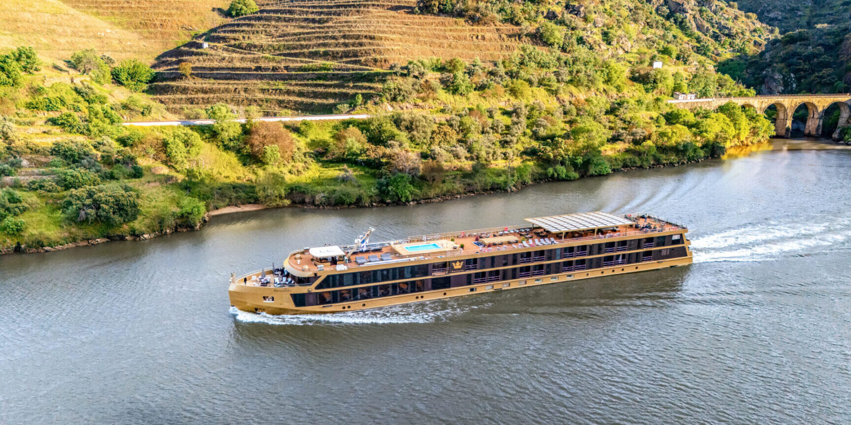 A large river cruise ship sails past terraced hillsides under bright daylight.